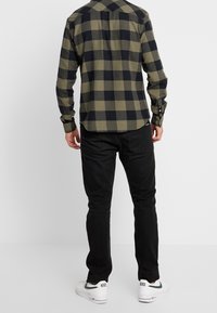 Camisa de franela en patrón de cuadros olivo y negro, con mangas de botón, combinada con jeans negros y zapatillas blancas con acentos verdes.