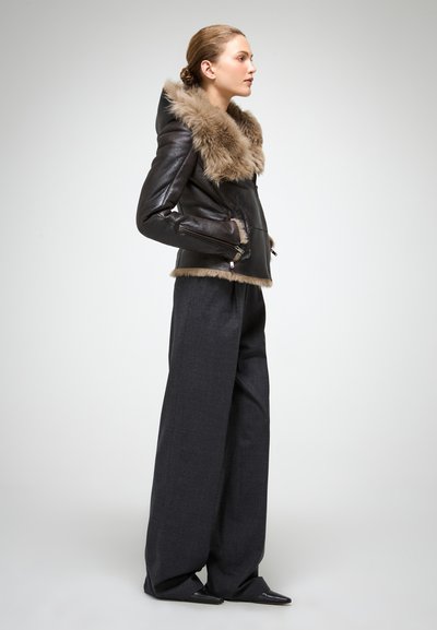 Braune Lederjacke mit einer Fellkapuze, ausgestattet mit Reißverschlusstaschen und einem kurzen Saum; kombiniert mit lockeren, dunkelgrauen Hosen und schwarzen High Heels.