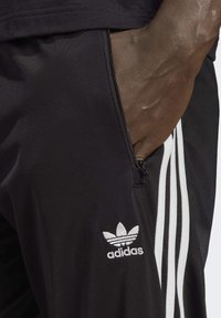 Pantalones atléticos negros hechos de un tejido suave. Presentan un diseño de tres rayas blancas y un logo de adidas bordado en el muslo izquierdo, incluye un bolsillo con cremallera.