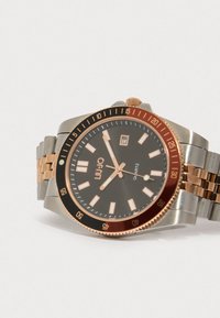 Ruostumattomasta teräksestä valmistettu rannekello mustalla kellotaululla, ruusukultaisilla yksityiskohdilla, valoa heijastavilla merkkivaloilla, päivämääränäytöllä ja kääntyvällä kehällä kahdenvärisessä metallirannekkeessa.