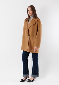 Cappotto in misto camel con ampi rever, due tasche anteriori e un fit rilassato, abbinato a jeans blu scuro e scarpe nere a punta.