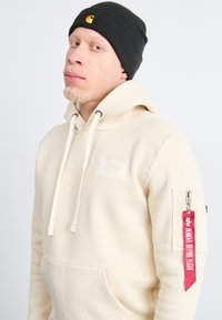 Hoodie couleur crème avec une poche avant, capuche à cordon de serrage et un patch. Comprend une poche zippée et une étiquette rouge avec "ENLEVER AVANT LE VOL." Bonnet noir.