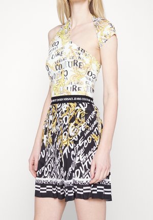 Femme portant un haut asymétrique sans manches et une jupe plissée avec un motif baroque noir, blanc et or et le texte « Versace Jeans Couture ».