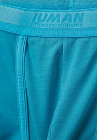 Turkosa joggers med en mjuk textur, med ett elastiskt midjeband präglat med "HUMAN." Designen inkluderar sidfickor och diskreta sömdetaljer.