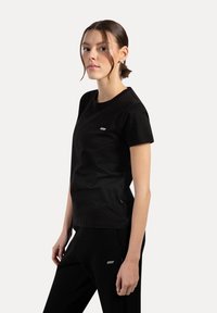 Camiseta de algodón negra con cuello redondo, mangas cortas y un pequeño parche de logotipo en el pecho para branding. Textura suave, ajuste regular.