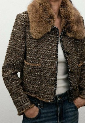 Bruine getextureerde tweed jas met grote bontkraag gedragen over een witte top en blauwe spijkerbroek, handen nonchalant in de zakken.