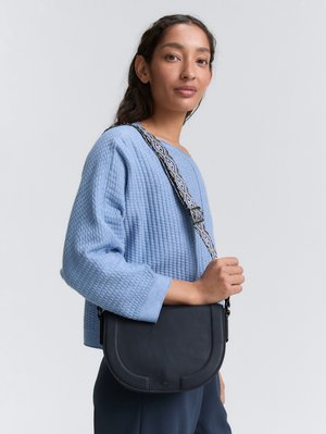 Marinefarbene Ledertasche in Kreuzbody-Form mit einer geschwungenen Silhouette, ausgestattet mit einem gemusterten Riemen und einer strukturierten Oberfläche. Die Tasche wird über einem hellblauen, strukturierten Pullover getragen.