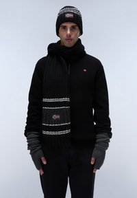 Sort strikket sweater, matchende tørklæde med hvide striber og flagdetalje, sort beanie med mønstrede accenter, grå fingerløse handsker.