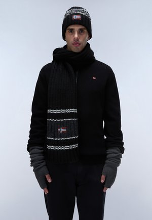 Sort strikket sweater, matchende tørklæde med hvide striber og flagdetalje, sort beanie med mønstrede accenter, grå fingerløse handsker.