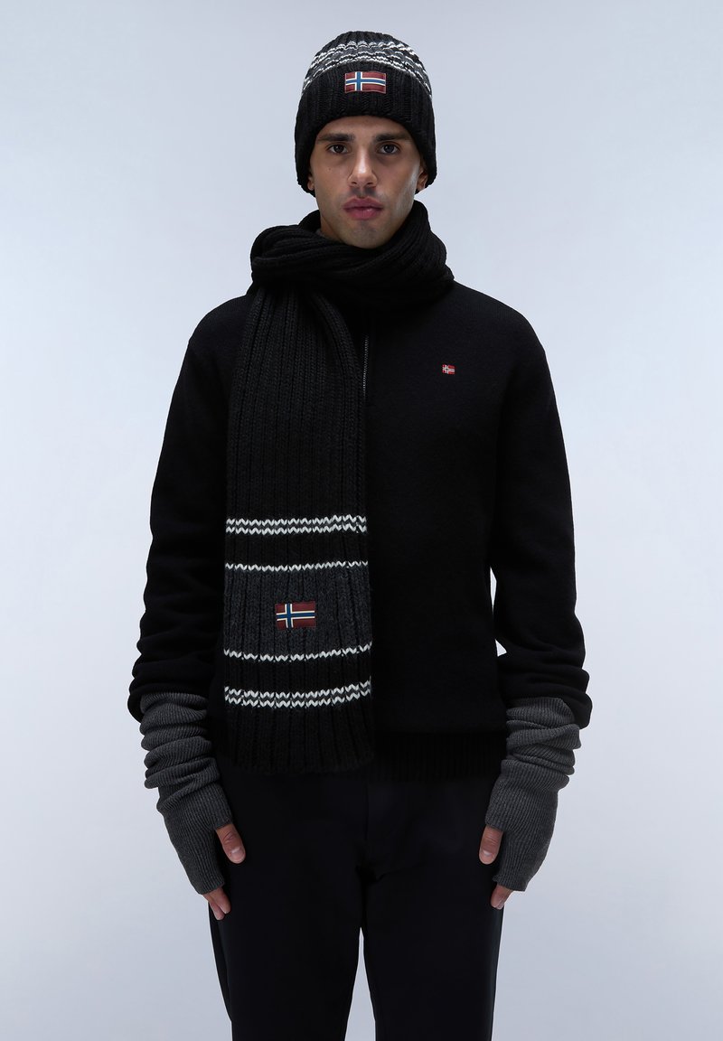 Sort strikket sweater, matchende tørklæde med hvide striber og flagdetalje, sort beanie med mønstrede accenter, grå fingerløse handsker.