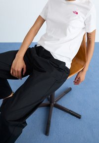 Witte t-shirt met korte mouwen en een klein roze logo op de borst, gecombineerd met losse zwarte broek, zittend op een houten stoel tegen een blauwe achtergrond.