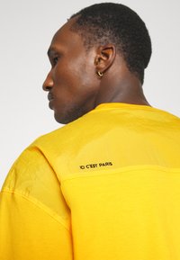 Gul T-shirt med en tekstureret, blank overdel og teksten "ICI C'EST PARIS" trykt i sort på bagsiden.