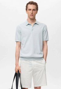 Polo de culoare albastră deschis, cu mânecă scurtă, dintr-un material neted, asortat cu pantaloni scurți albi. Modelul ține o geantă tote neagră, stând drept în fața unui fundal simplu.