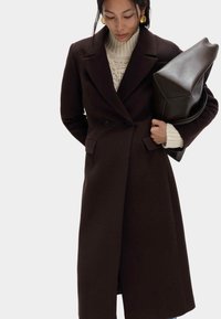 Długi, ciemnobrązowy płaszcz dwurzędowy z dużymi klapami, bocznymi kieszeniami i zbudowanymi ramionami. Modelka ma na sobie również kremowy sweter i trzyma torbę.