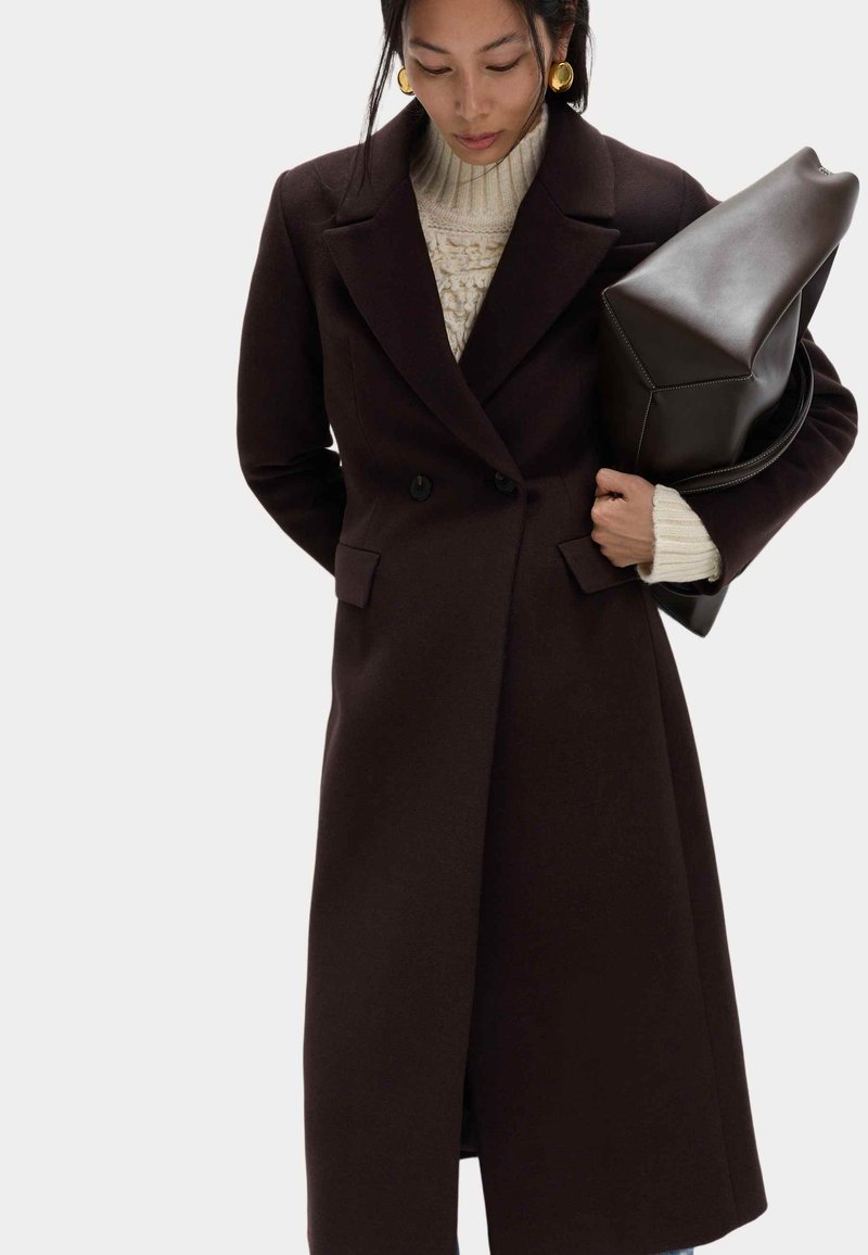 Długi, ciemnobrązowy płaszcz dwurzędowy z dużymi klapami, bocznymi kieszeniami i zbudowanymi ramionami. Modelka ma na sobie również kremowy sweter i trzyma torbę.