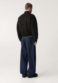 Giubbotto bomber in suede nero con vita a costine, abbinato a jeans di denim scuri e dalla vestibilità ampia. La vista posteriore mette in evidenza la silhouette e le cuciture.