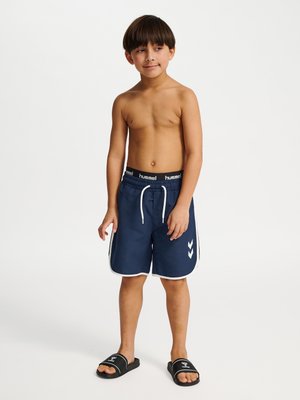 Jongen draagt marineblauwe zwemshort met witte bies en zwarte slippers, staat tegen een lichte achtergrond en kijkt iets naar rechts.