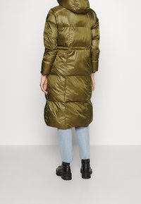Cappotto imbottito verde oliva con cappuccio, caratterizzato da una texture trapuntata e un design di lunghezza media. Abbinato a jeans azzurri chiari e stivali neri.