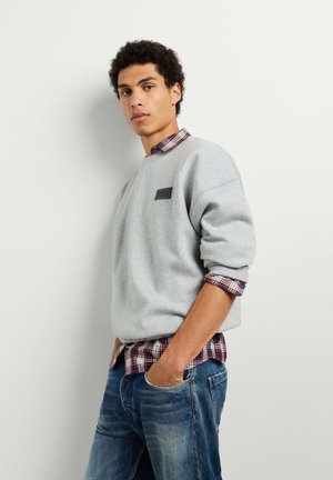 Junger Mann steht mit den Händen in den Taschen, trägt einen grauen Sweatshirt über einem roten karierten Hemd und blaue Jeans vor hellem einfarbigem Hintergrund.