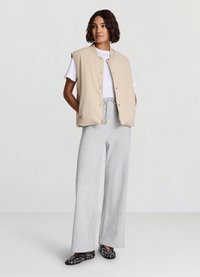 Beige ärmellose Steppweste über einem weißen T-Shirt, kombiniert mit grauer Jogginghose und schwarzen Schuhen mit runden Ausschnitten. Glatte Textur, lockere Passform.