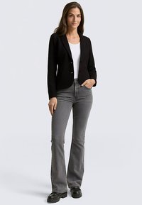 Blazer nero aderente sopra una camicia bianca, abbinato a jeans grigi svasati e stivali neri. Tessuto liscio, design sartoriale e due bottoni.