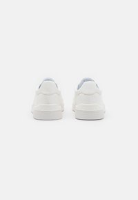 Zapatillas blancas de corte bajo con parte superior de lona texturizada y suela de goma. Logo sutil en el talón, punta redondeada, diseño minimalista.