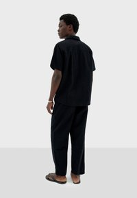 Schwarzes kurzärmeliges Hemd und lockere Hose, strukturierter Stoff, kragenloses Design, kombiniert mit Flip-Flops. Rückansicht, minimalistischer Stil.