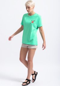 Mintgroene T-shirt met korte mouwen met een rode krabafbeelding op de voorkant, gecombineerd met beige shorts en zwarte sandalen.