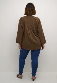 Brun langærmet skjorte med buet kant og rygpanel, parret med blå skinny jeans og iført sorte hæle.