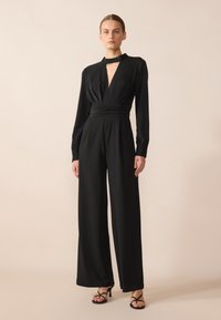 Combinaison noire à manches longues, décolleté en V profond, tissu froncé à la taille et design à jambes larges. Portée avec des talons hauts noirs à lanières.