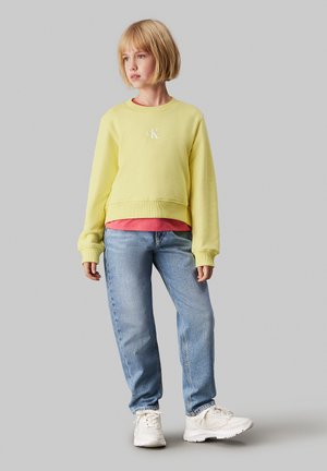 Sudadera amarilla corta con logotipo blanco, combinada con una camiseta roja, pantalones vaqueros azules y zapatillas blancas. Tela suave, corte holgado.