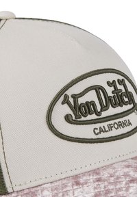 Casquette de baseball beige Von Dutch avec logo brodé vert et le mot "California" sur le panneau avant, posée sur une surface texturée rose.