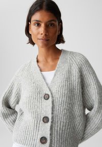 Donna con capelli corti scuri che indossa un cardigan spesso grigio chiaro con tre grandi bottoni scuri sopra una maglietta bianca.