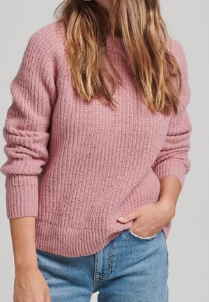 Femme portant un pull en maille rose texturé avec les manches retroussées et un jean bleu clair, la main posée négligemment dans la poche, fond neutre.