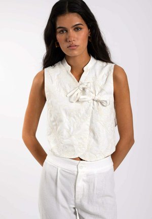 Top blanco sin mangas con cuello mao, que presenta un detalle de lazo y bordado floral en un tono beige claro. Combinado con pantalones de lino blancos.