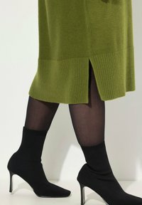 Robe tricotée olive jusqu'aux genoux avec ourlet côtelé et fente latérale, associée à des chaussettes noires ajustées jusqu'à la cheville et des escarpins à bout pointu.