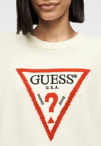 Κοντινό πλάνο ενός ατόμου που φοράει εκρού πουλόβερ με κόκκινο τρίγωνο λογότυπο που γράφει "GUESS U.S.A. Washed Jeans".