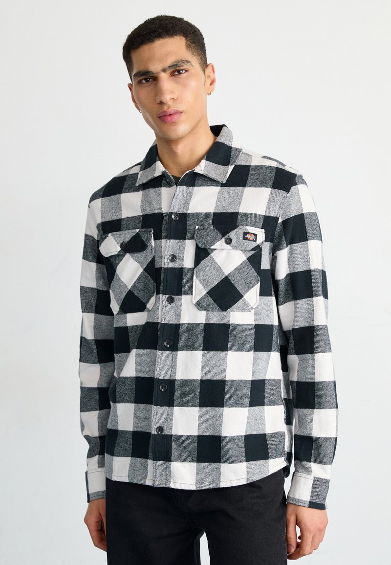 Camisa de franela a cuadros en blanco y negro con dos bolsillos en el pecho, cierre de botones y mangas largas, llevada sobre unos jeans oscuros.