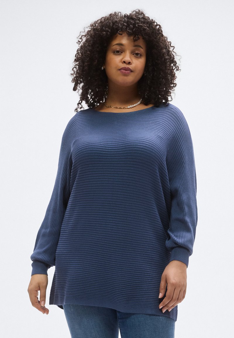 Μπλε ναυτικό ριγέ πουλόβερ με χαλαρή εφαρμογή, μακριά μανίκια και φαρδύ λαιμόκοψη, συνδυασμένο με ανοιχτόχρωμο τζιν. Ορατή υφή υφάσματος.