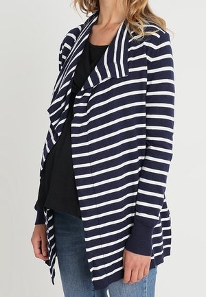 Woman wearing a navy blue and white striped open-front cardigan over a black top and blue jeans against a plain background.