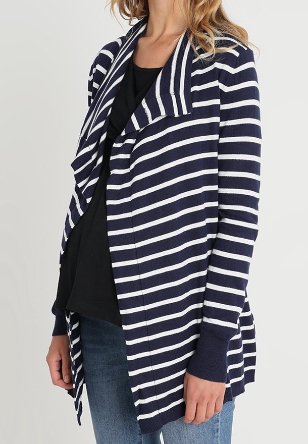 Femme portant un cardigan ouvert à rayures bleu marine et blanc sur un top noir et un jeans bleu, sur un fond uni.