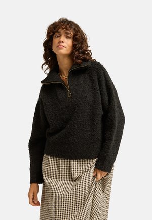 Donna con capelli ricci che indossa un maglione nero con zip e una gonna a quadri beige e nera, in piedi davanti a uno sfondo semplice.