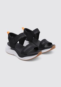 Sandalias negras con correas de tela anchas y cierres ajustables. Presentan una suela blanca texturizada y un pequeño acento naranja en el talón.