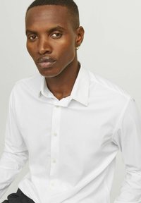 Camicia bianca in cotone con colletto strutturato, maniche lunghe e una texture liscia. Il modello ha i capelli corti e indossa un delicato orecchino a cerchio.
