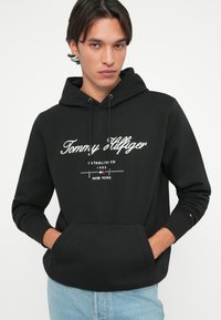 Black hooded sweatshirt with white text featuring "Tommy Hilfiger," a kangaroo pocket, and ribbed cuffs. Made of soft fabric, standard fit.