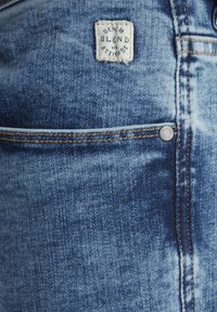 Plave traperice s svijetlim pranjem i kontrastnim narančastim šavovima. Uključuje oznaku s natpisom "DENIM BLEND" i metalnu kopču.