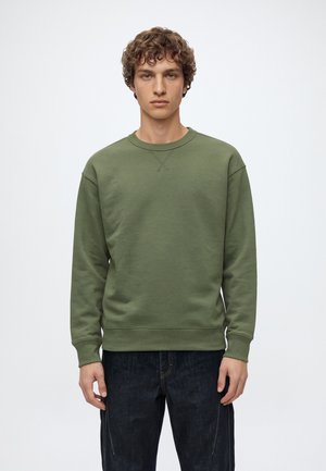 Un jeune homme porte un sweat-shirt uni vert olive à col rond et un jean foncé sur un fond clair de studio.