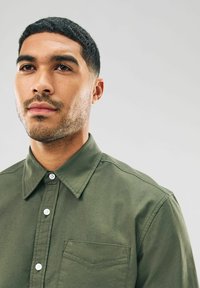 Chemise boutonnée olive en coton, dotée d'un col pointu, d'une poche frontale et de boutons blancs, avec une texture lisse.