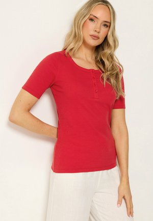 Vrouw met lang blond haar die een rood kort mouw knoopjes overhemd en witte broek draagt, staand tegen een effen lichte achtergrond.