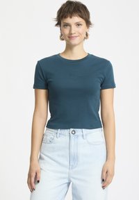 Tealfarbene kurzärmelige Baumwoll-T-Shirt mit Rundhalsausschnitt und dezenten Logodetails, kombiniert mit hochtaillierten hellblauen Jeans.